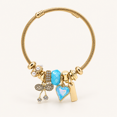 Gold-tone blue charm bracelet with crystal bow, pearl accents and heart pendant on a cream background.