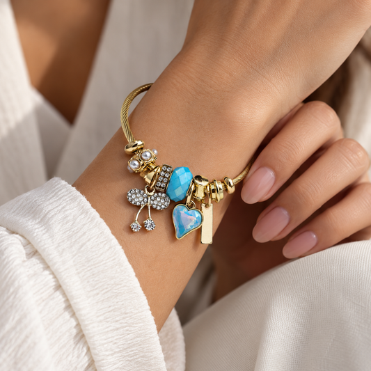 Close-up of gold cable charm bracelet with blue heart, turquoise-blue bead and sparkling crystal bow on a woman's wrist.