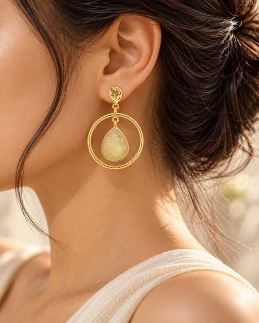Close-up of textured gold circular hoop earrings with delicate gemstone-style teardrop charms.