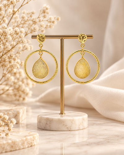 ANOVRA Golden Teardrop Hoops displayed on a black jewellery stand with minimal floral props and a cream background.